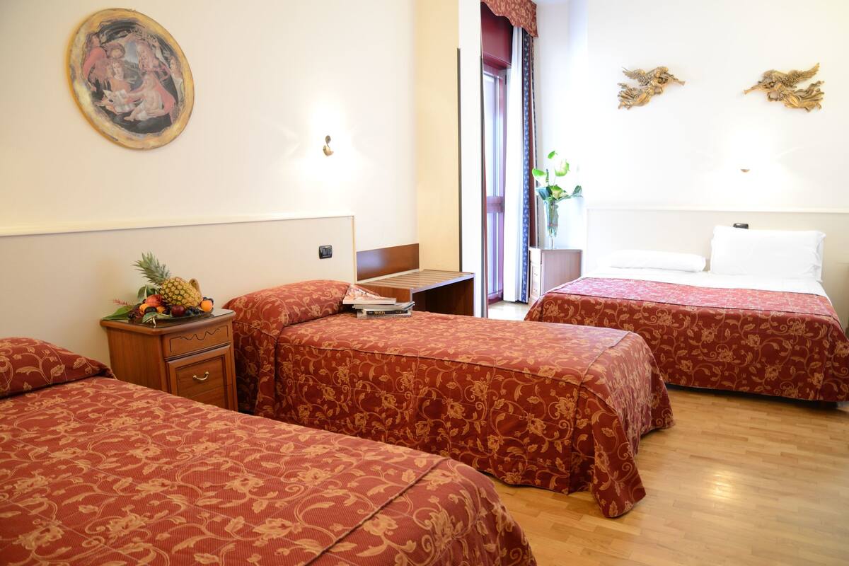 A hotel room with two beds, a wooden nightstand with a fruit bowl, a painting on the wall, and a window with curtains.