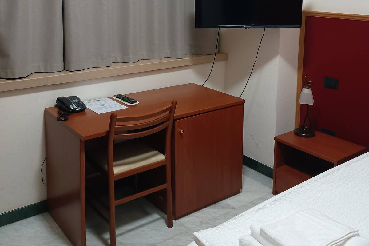 A hotel room with a wooden desk, a chair, a television mounted on the wall, a bedside table with a lamp, and a bed with a folded white towel.
