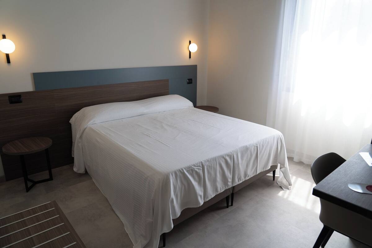 A neatly made bed in a hotel room with a wooden headboard, two wall-mounted lights, a small round table, and a chair.