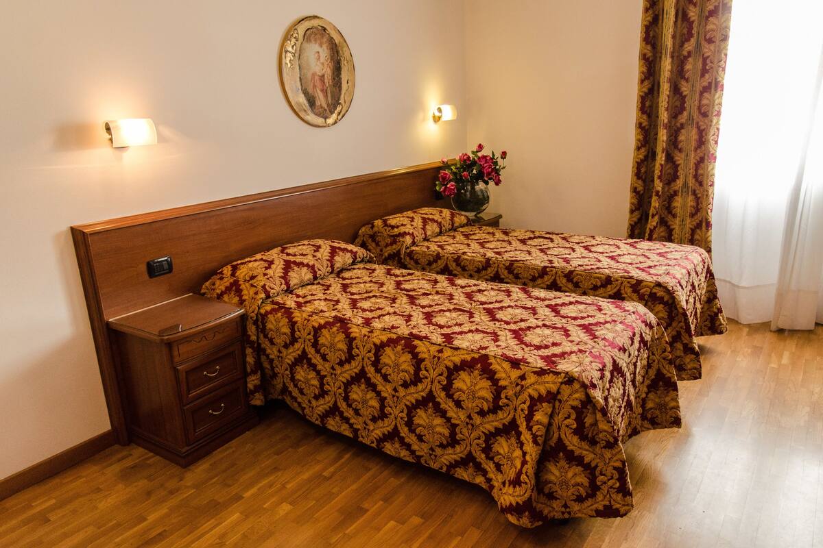 A room with two beds, wooden headboard, nightstands, a wall-mounted light, a framed picture, and a curtain.
