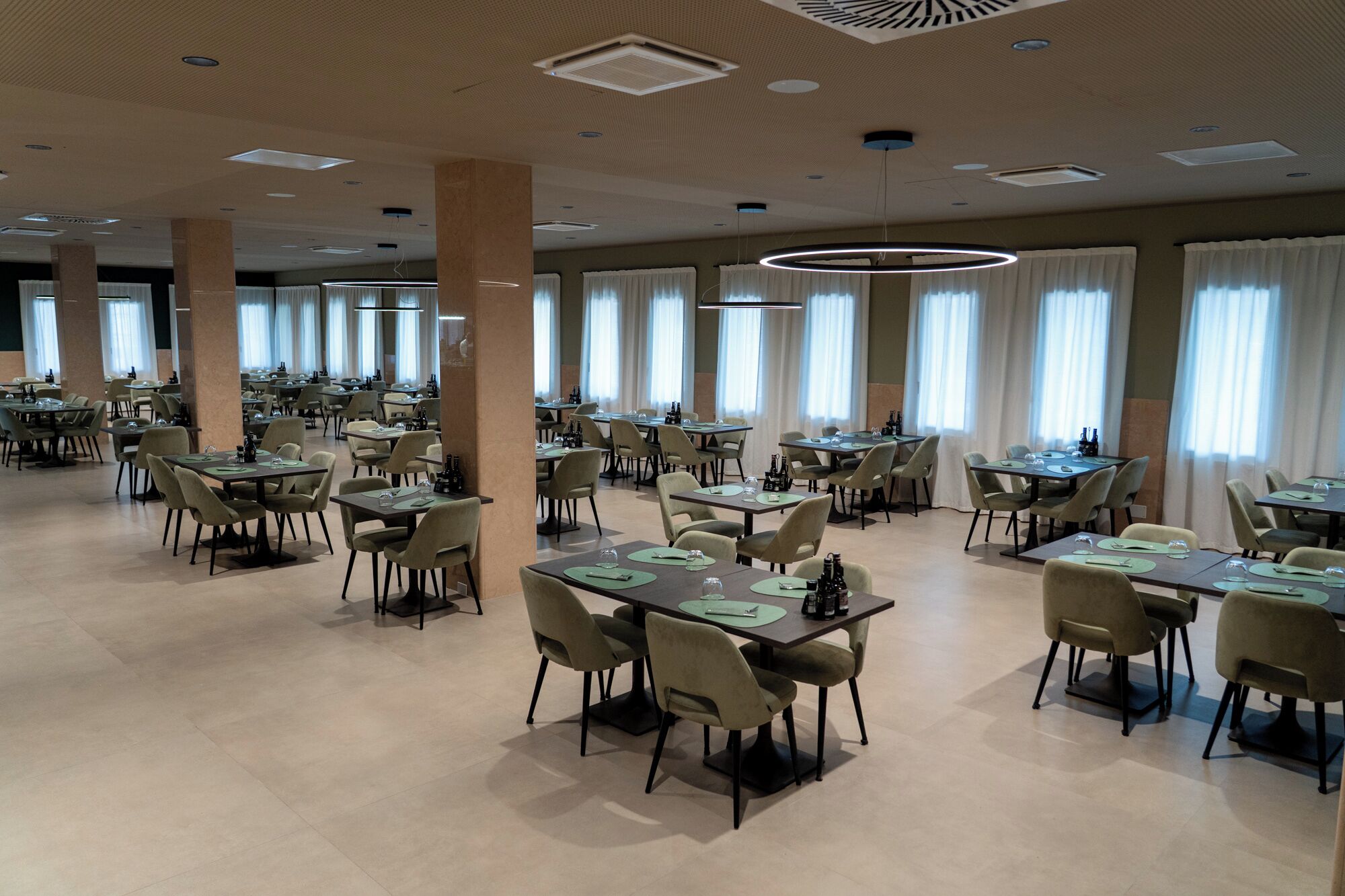 Un vaste espace dédié à la salle à manger, avec de nombreuses tables et chaises, de grandes fenêtres agrémentées de rideaux et un design moderne au niveau du plafond.