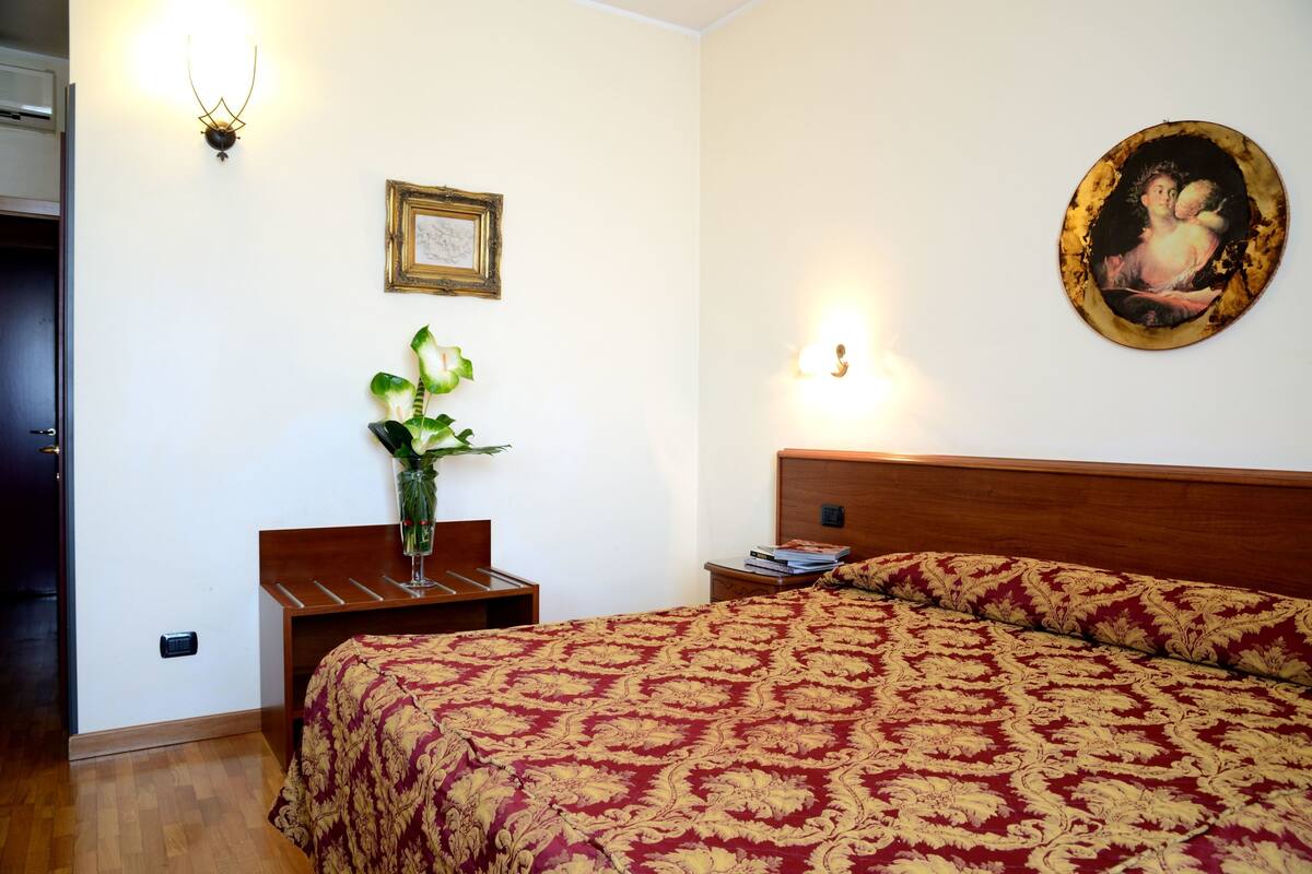 A bedroom with a wooden headboard, a large bed with a red and gold patterned bedspread, a nightstand with a vase of flowers, a framed picture on the wall, and a wall-mounted light fixture.