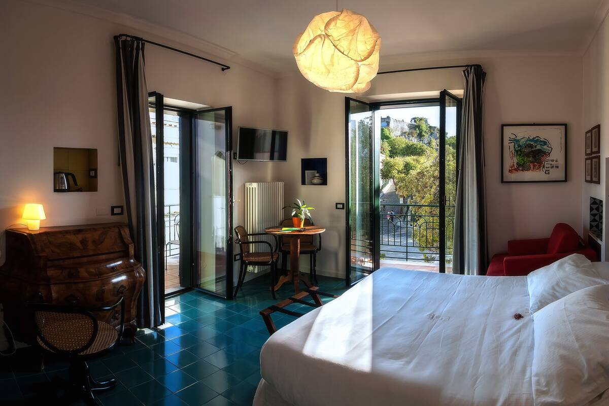 Une chambre d’hôtel comprenant un lit, un bureau avec une chaise, une table avec un pot de plante, une chaise rouge, une grande fenêtre donnant sur l’extérieur et un luminaire suspendu.