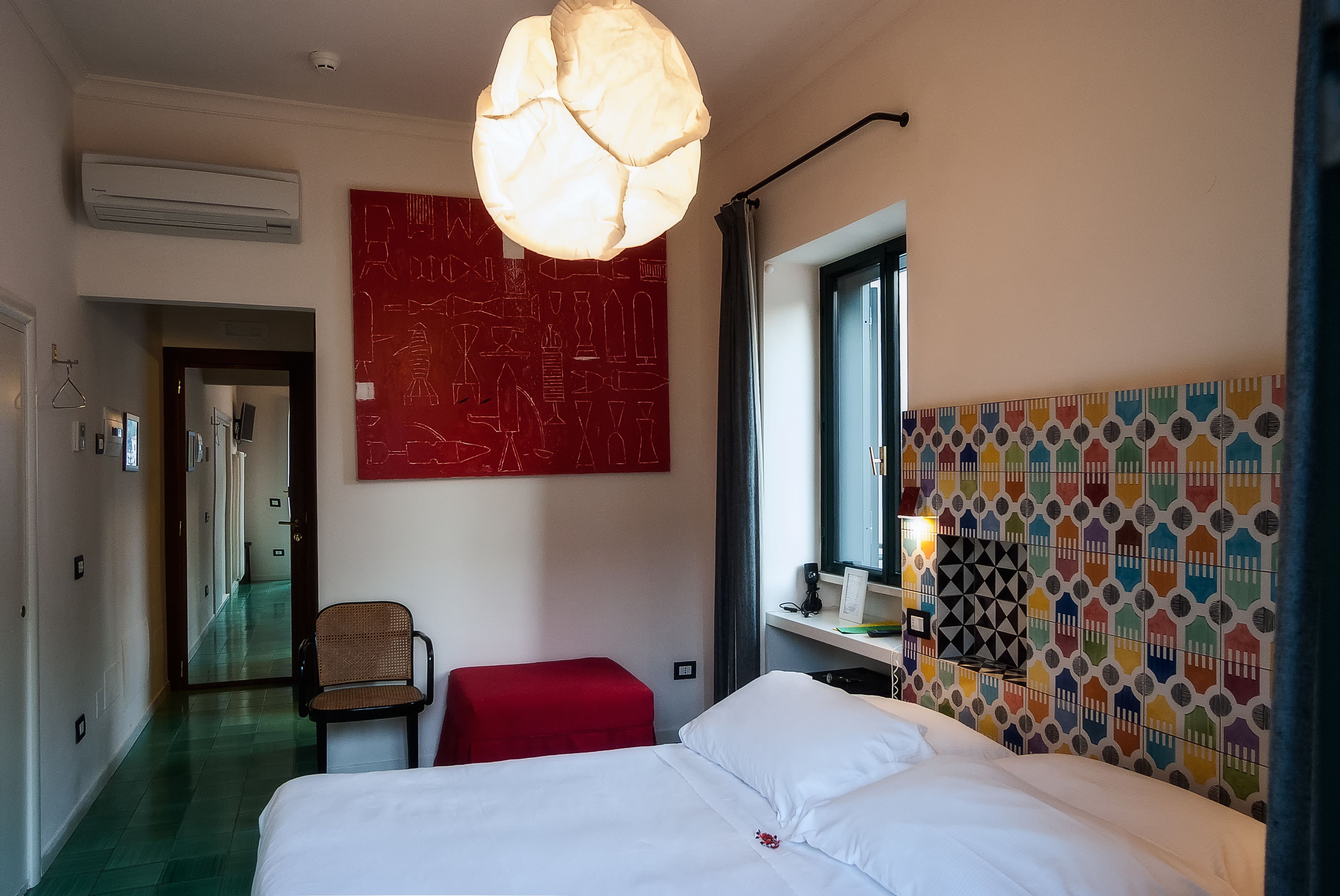 Een hotelkamer met een bed, een rode stoel, een rood poefje, een kunstwerk aan de muur, een behang met patroon en een raam met gordijnen.
