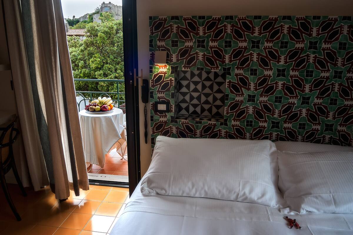 Une chambre d’hôtel avec un lit, une vue sur un balcon agrémenté d’une table et de fruits, et un mur orné d’un motif.