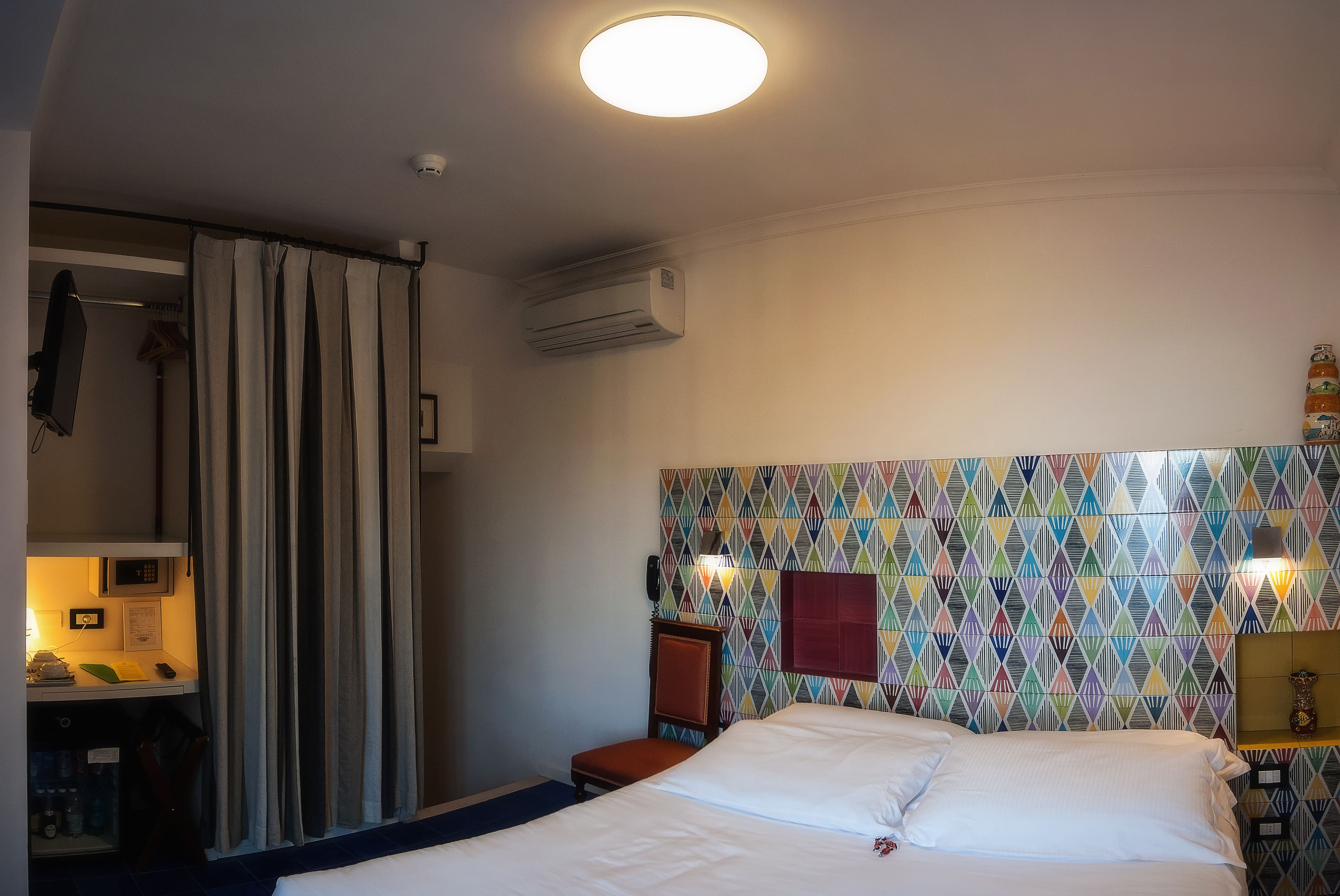 A hotel room with a bed, a chair, a desk with a lamp, and a colorful wall pattern.
