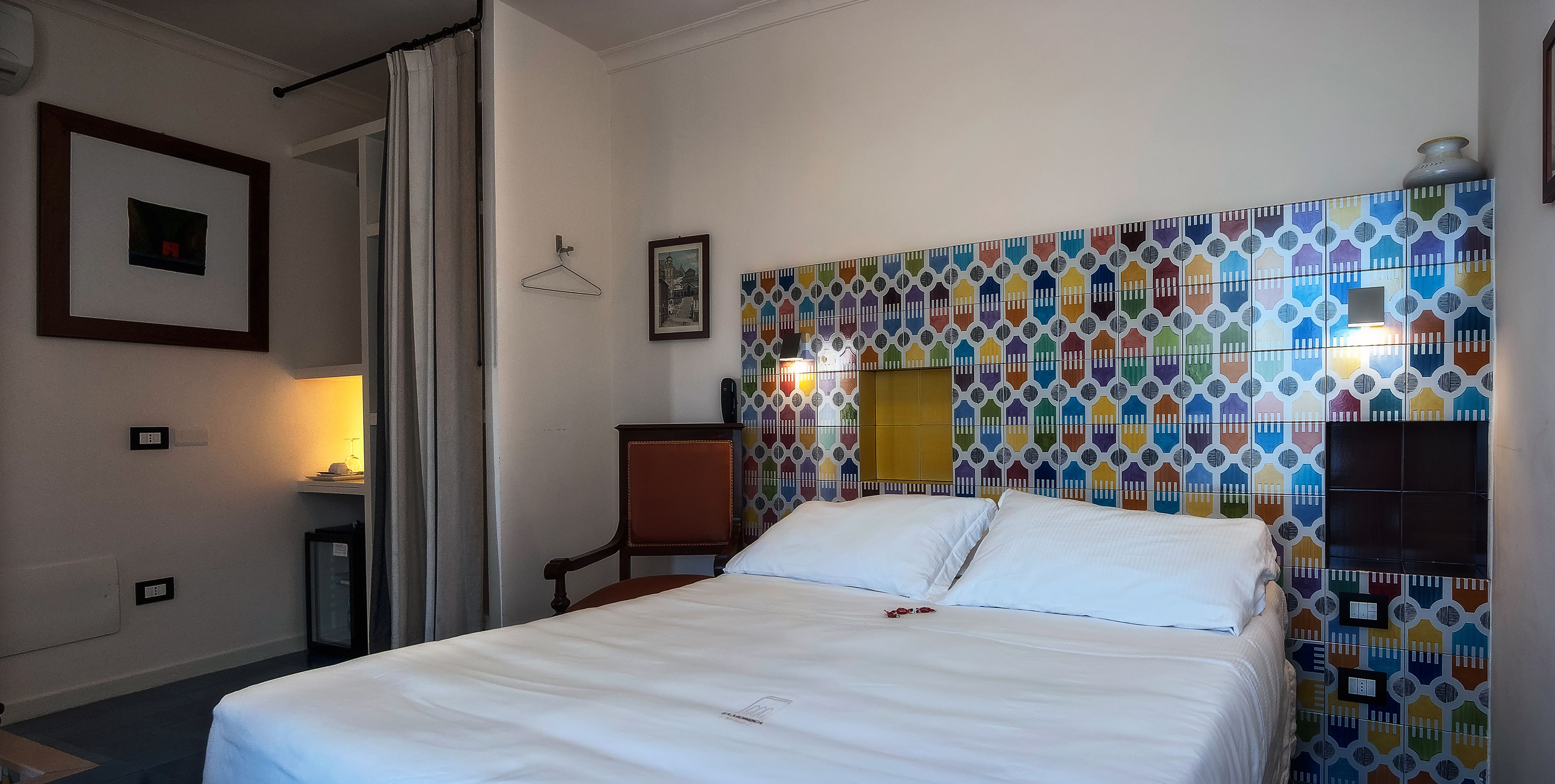 Habitación de hotel con cama, una silla, una mesita y una pared de azulejos coloridos.