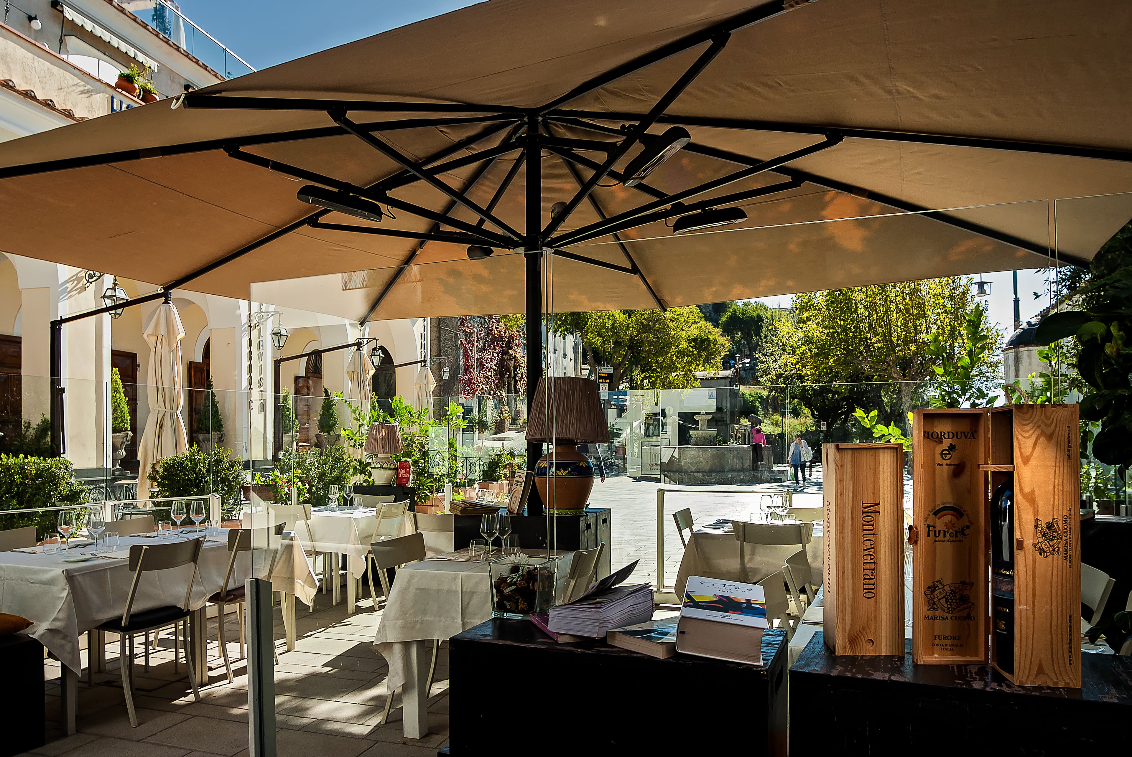 An outdoor dining area with tables and chairs under a large umbrella, surrounded by potted plants and a street view.