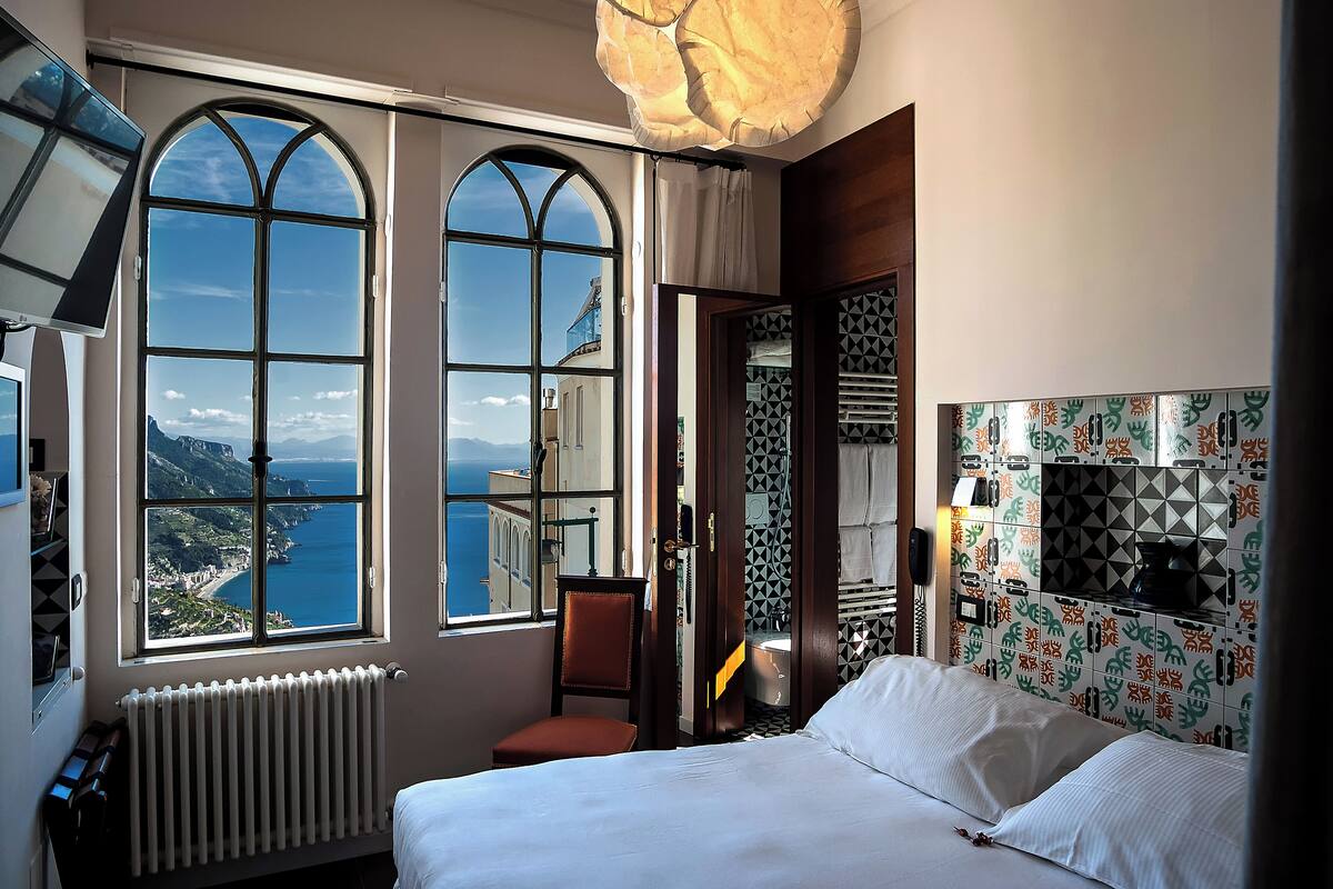 Une chambre d’hôtel dotée d’une grande fenêtre en arc offrant une vue panoramique, d’un lit, d’une chaise et d’une petite salle de bain aux carreaux décoratifs.