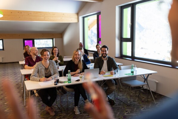 A group of people seated at tables in a room with large windows and a wooden ceiling.