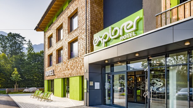 Een modern hotel met een groen-bruine gevel, een ruime glazen entree en een terras.