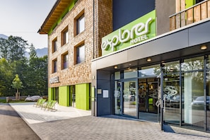 A modern hotel with a green and brown facade, large glass entrance, and outdoor seating area.