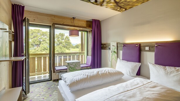 A hotel room with two beds, a desk, and a window with purple curtains.