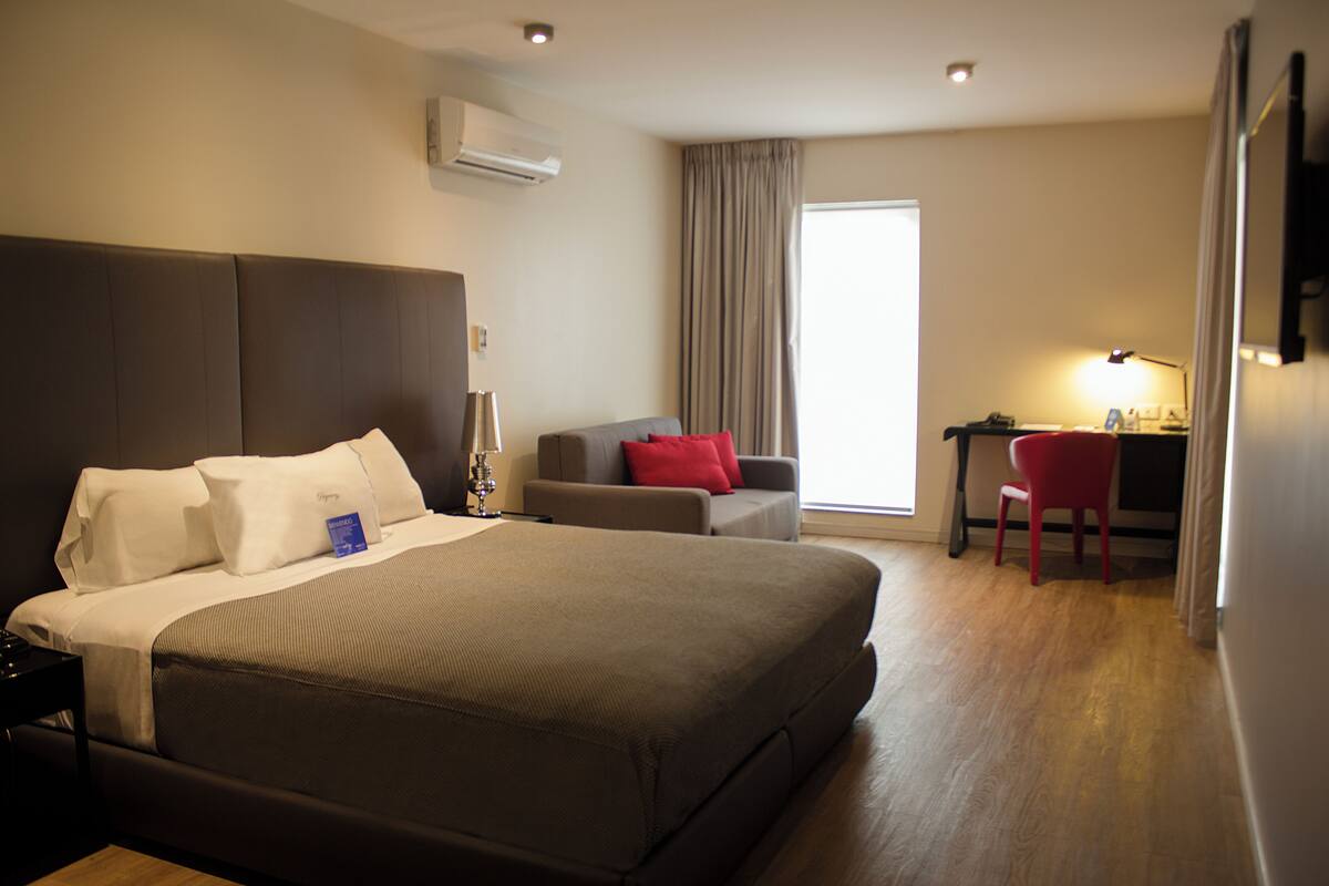 Una habitación de hotel con una cama grande, un escritorio, una silla y un ventanal con cortinas.