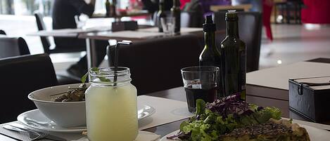A dining table with a meal consisting of a salad, a slice of quiche, and a glass of lemonade. There are also two bottles of wine and a glass of water on the table.