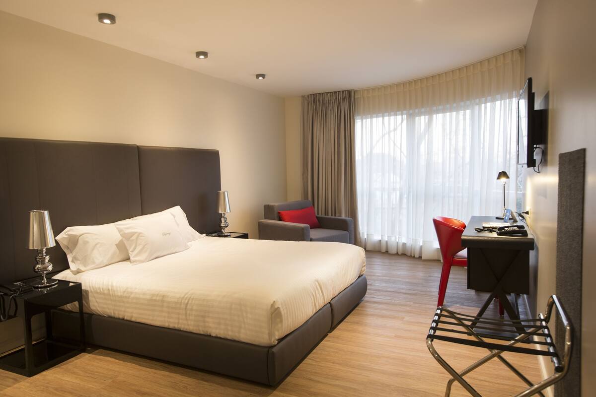 Una habitación de hotel moderna con una cama grande, un escritorio con silla, televisión y una lámpara.