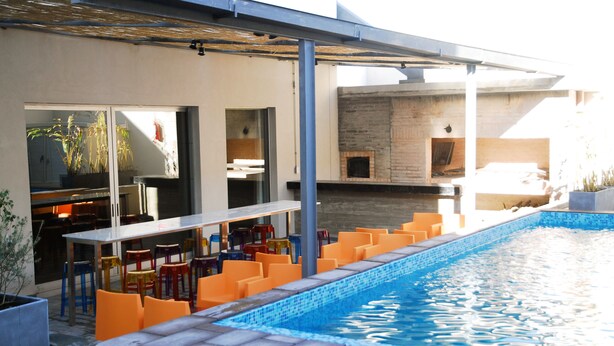 Zona de piscina con sillas naranjas y una mesa, chimenea de ladrillo y puerta de vidrio que da a una habitación.