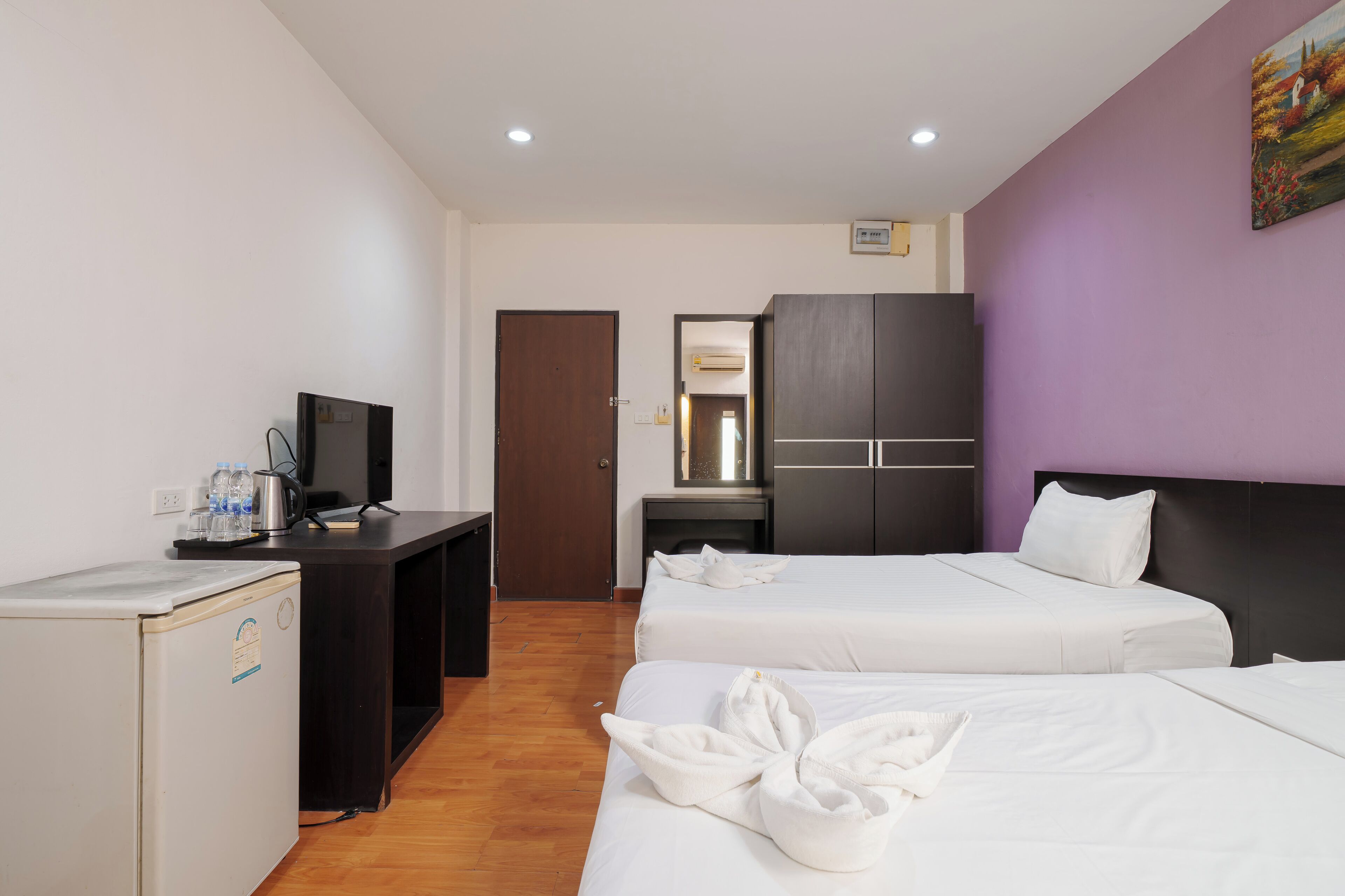 A hotel room with two beds, a desk with a computer, a refrigerator, and a wardrobe.