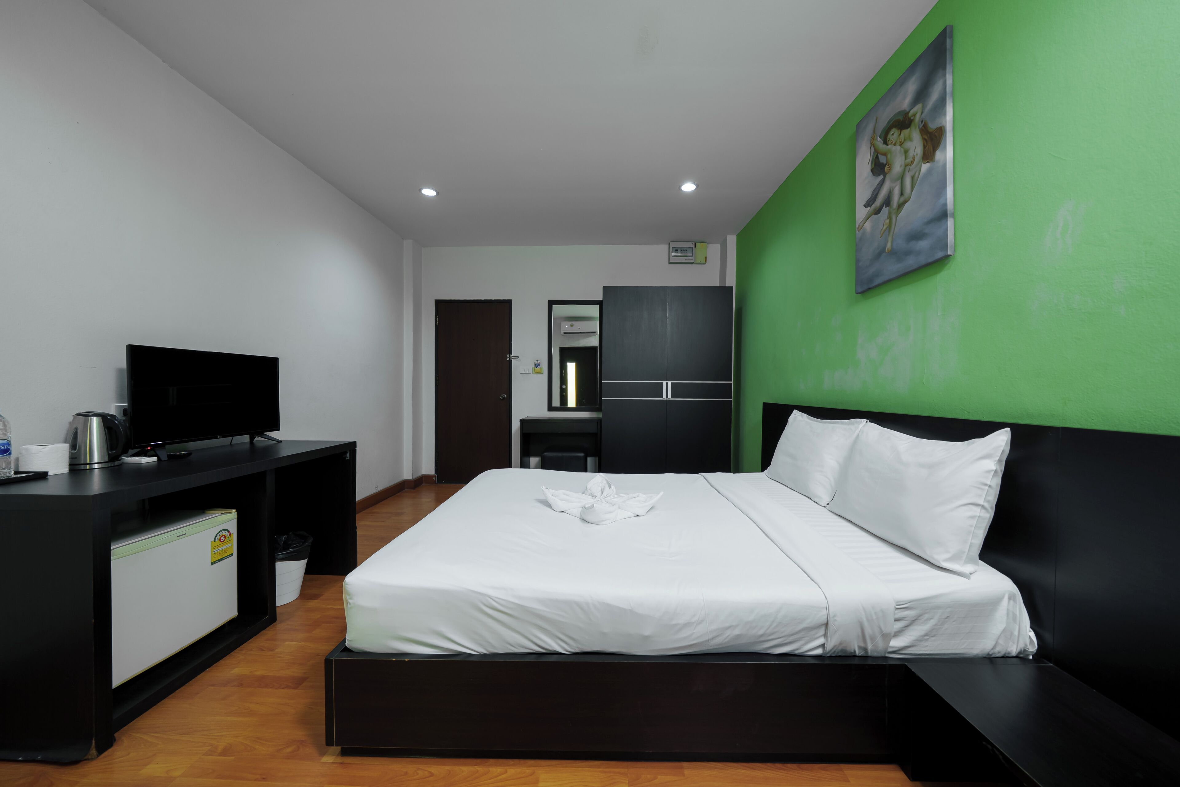 A modern hotel room with a bed, a desk with a television, a small refrigerator, and a green accent wall.