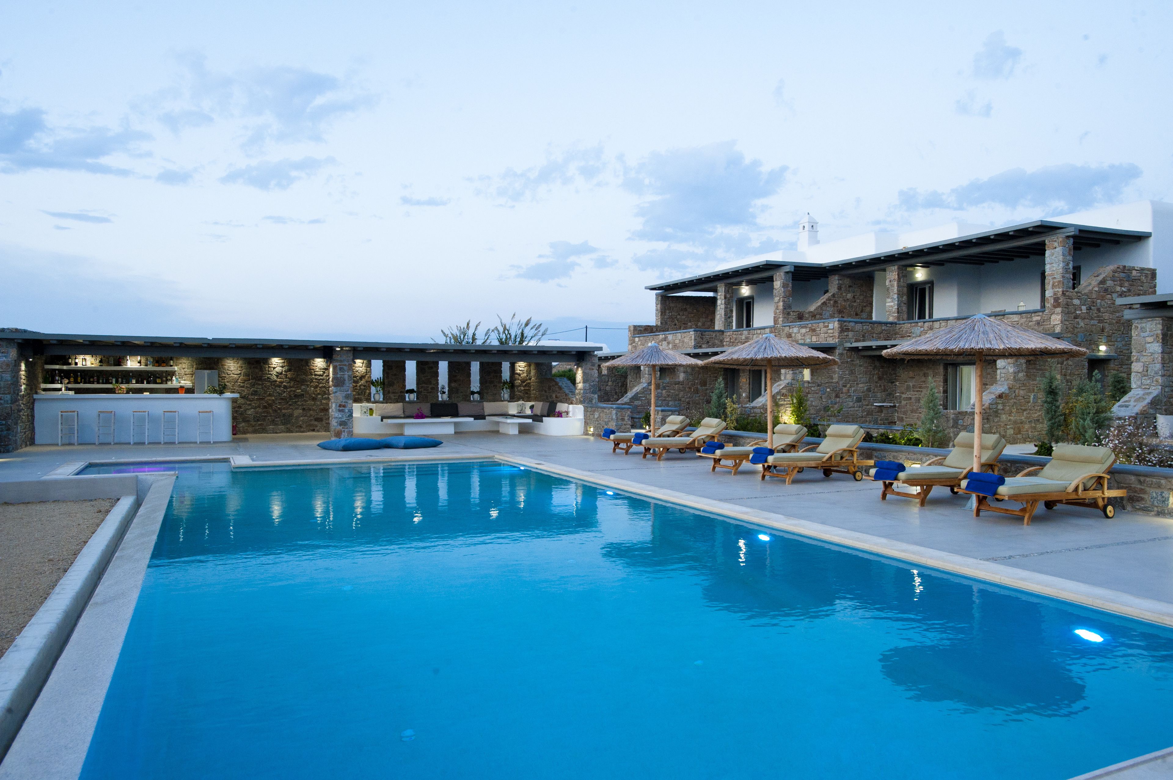Una piscina con sillones de descanso y sombrillas, un área de bar y un edificio de piedra con balcón.