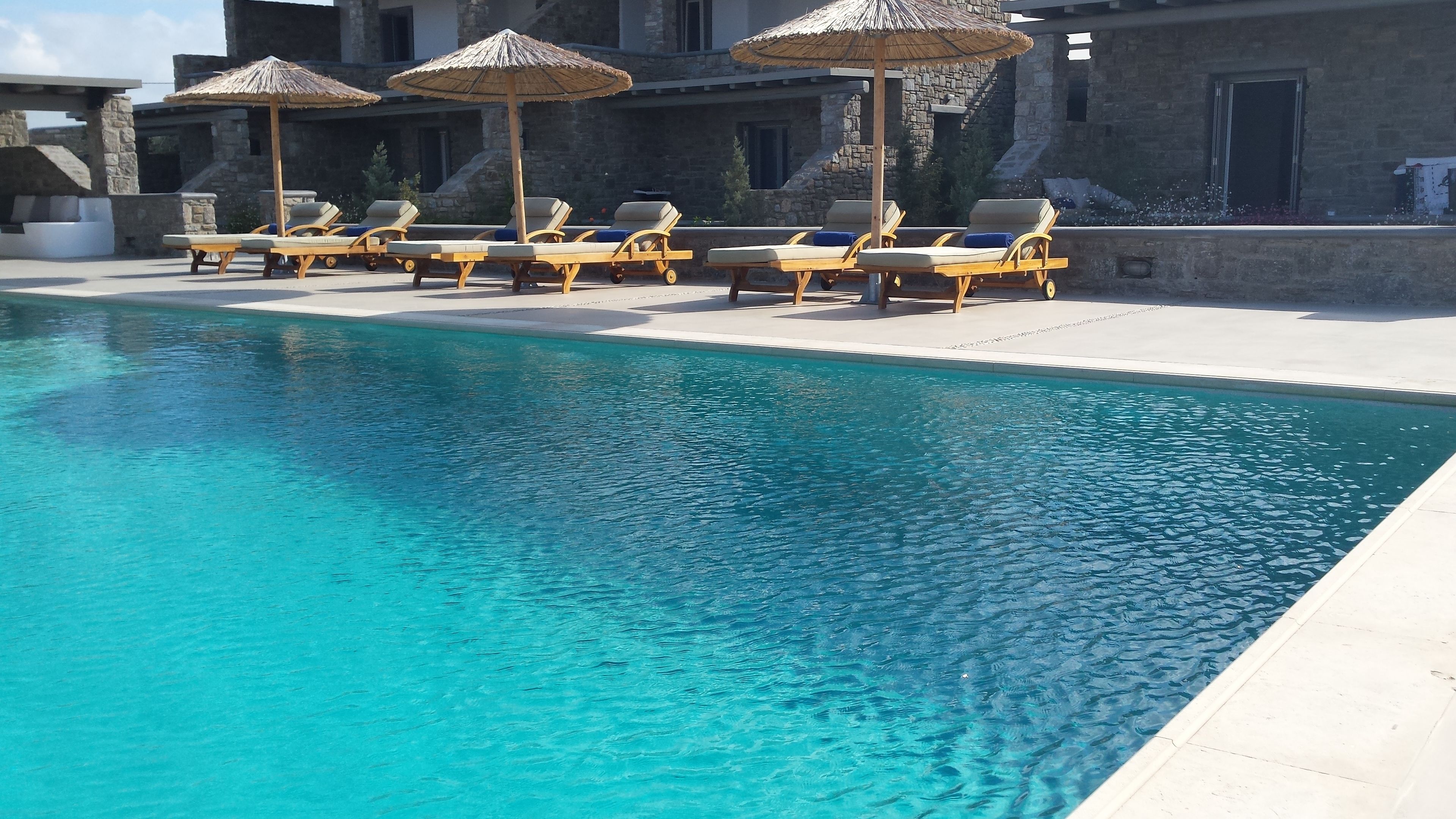 Un área de piscina con sillas de descanso y sombrillas, edificios de piedra al fondo y una piscina azul cristalina.