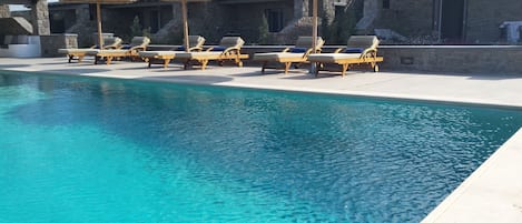Un área de piscina con sillas de descanso y sombrillas, edificios de piedra al fondo y una piscina azul cristalina.