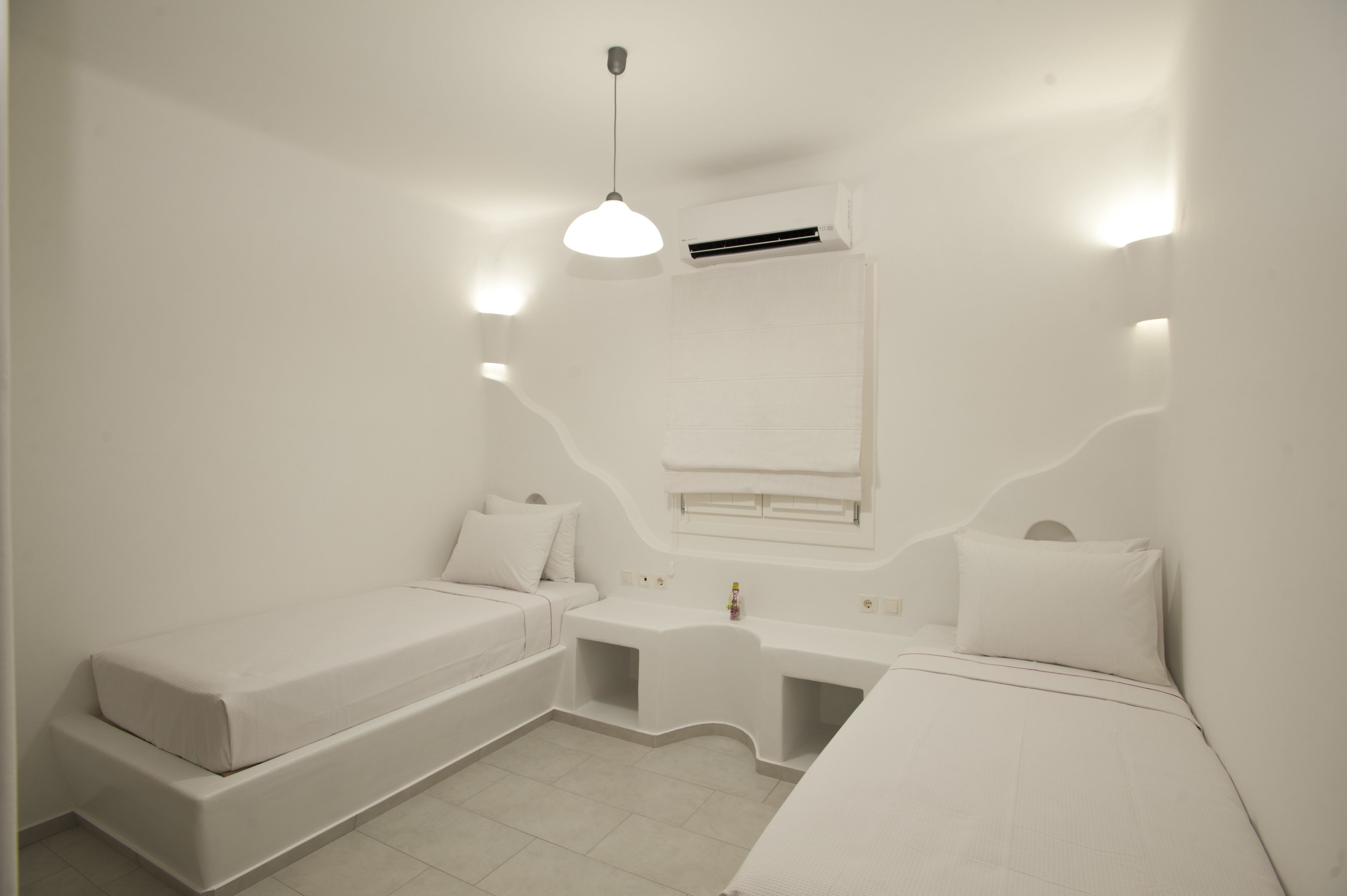 Un dormitorio minimalista y blanco, con una cama, un escritorio pequeño y una lámpara colgante.