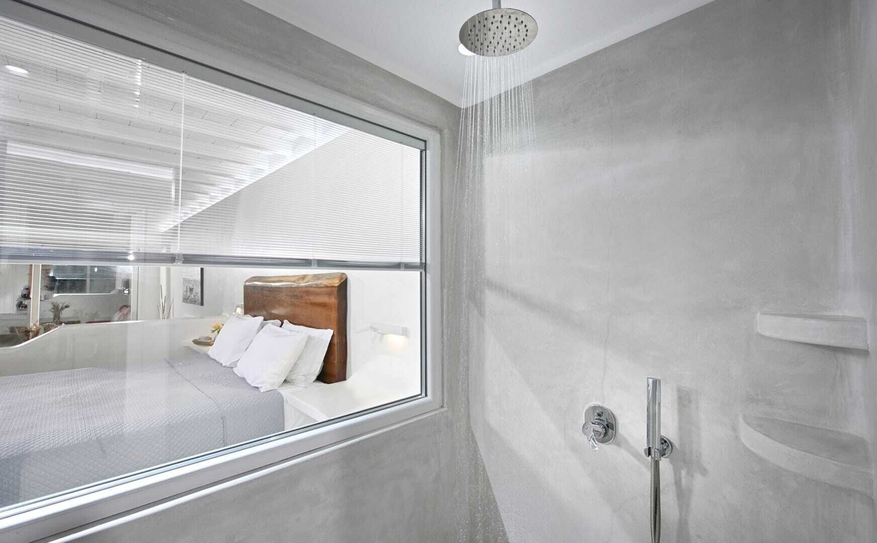 A modern bathroom with a shower, a large window, and a bed visible through the window.