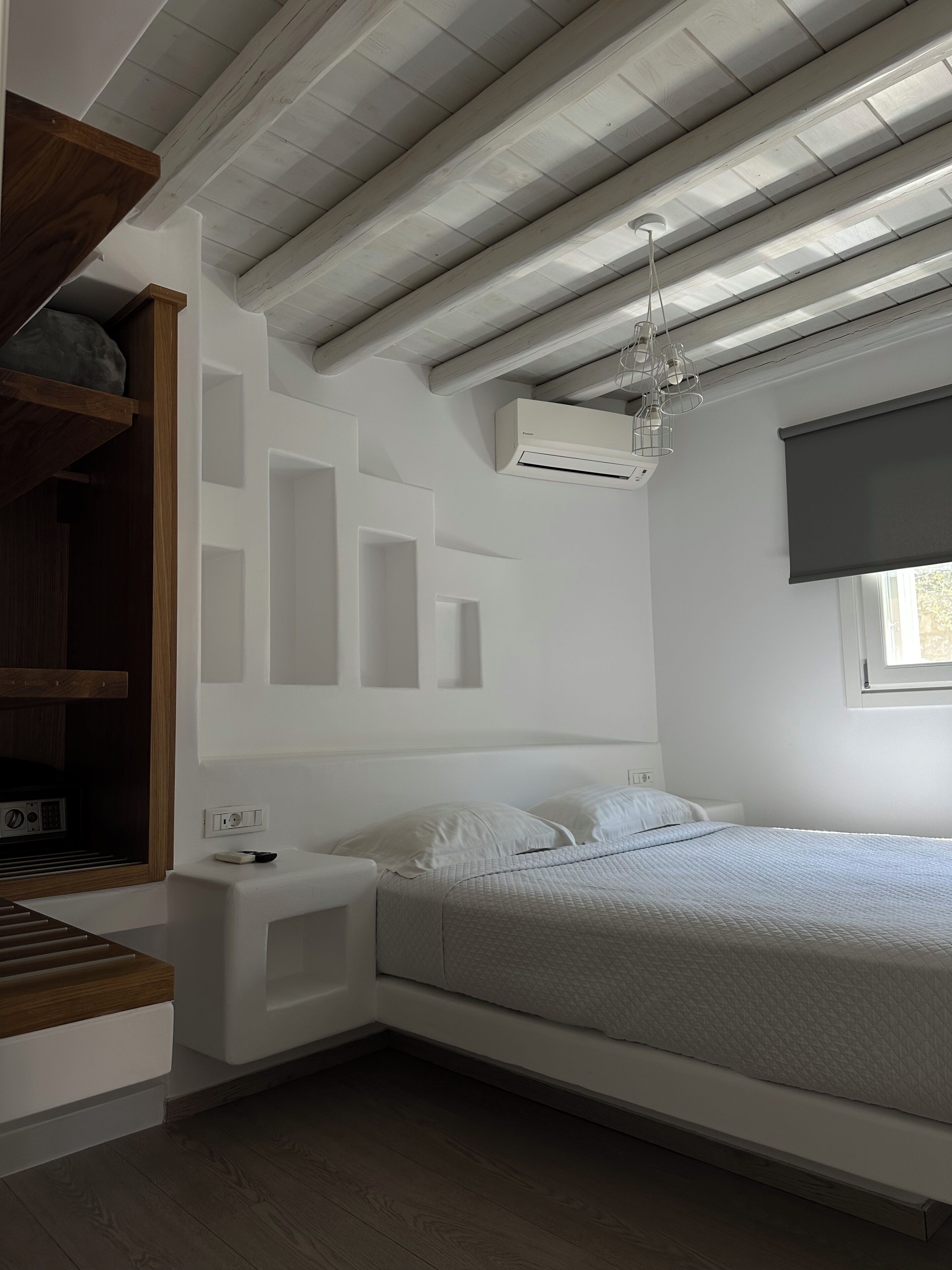 Un dormitorio con cama, mesita de noche, estanterías empotradas, unidad de aire acondicionado, ventana con persiana y techo de madera.