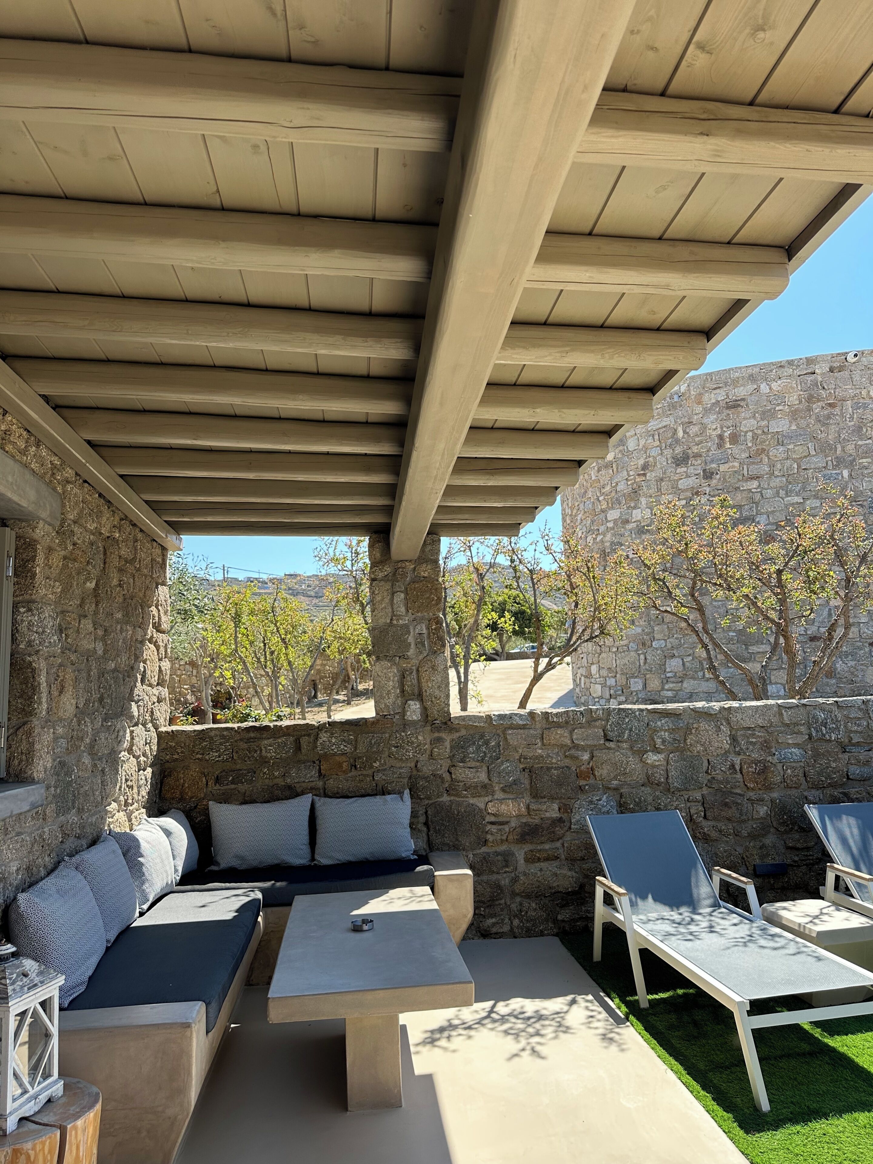 Un espacio de estar al aire libre techado con paredes de piedra, una mesa de piedra y sillones de descanso.