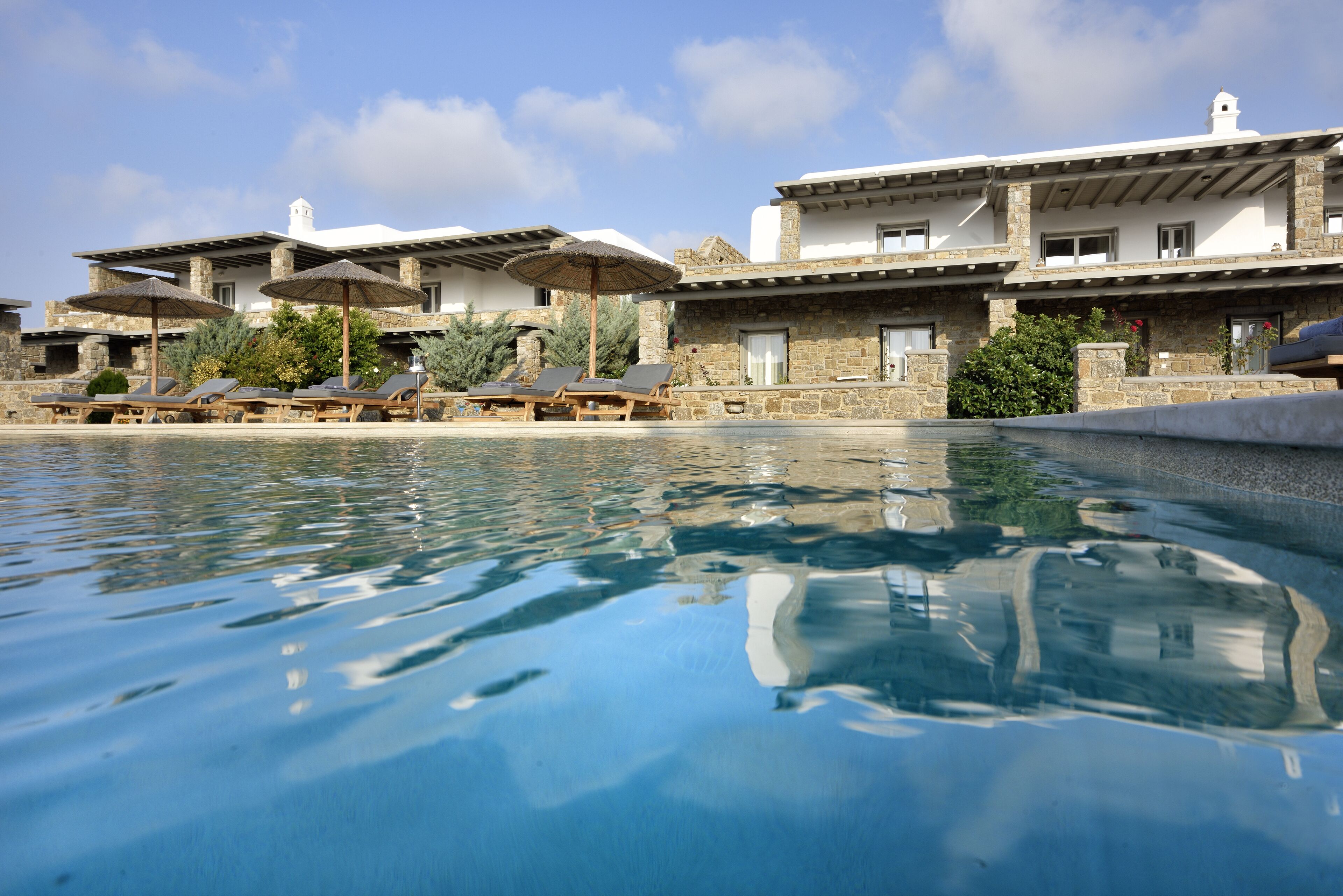 Área de piscina com espreguiçadeiras e guarda-sóis, edifícios de pedra com varandas e céu claro.