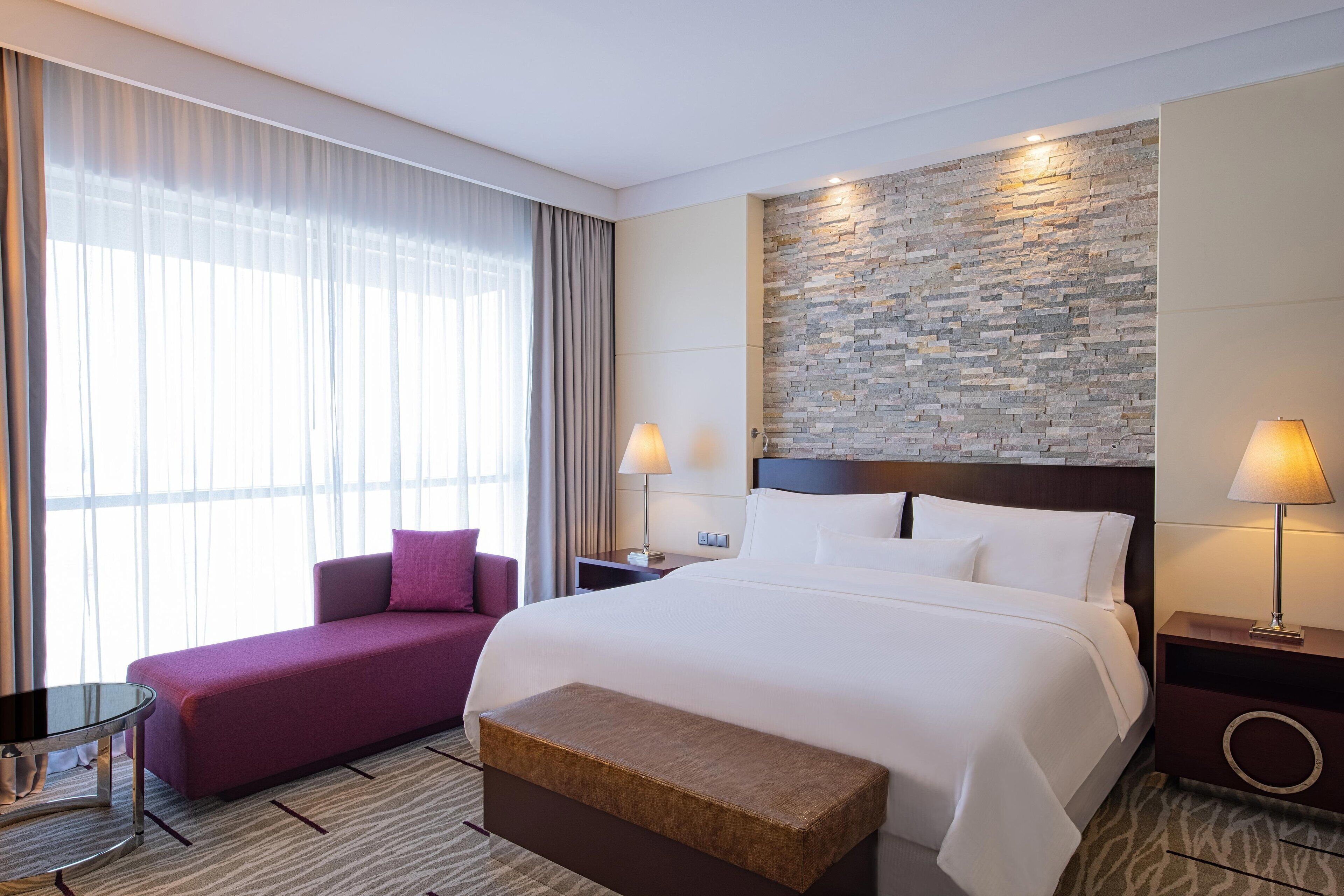 A hotel room with a large bed, a sofa, a bench, a nightstand, and a wall with a stone pattern.