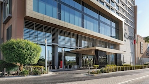 Un hotel moderno con fachada de vidrio, una entrada destacada y una zona ajardinada con árboles y arbustos.