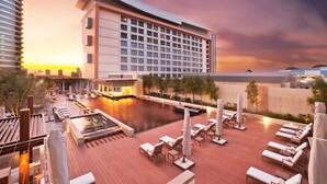 Un área de piscina en la azotea con sillones de descanso y sombrillas, un gran edificio de hotel y un atardecer al fondo.