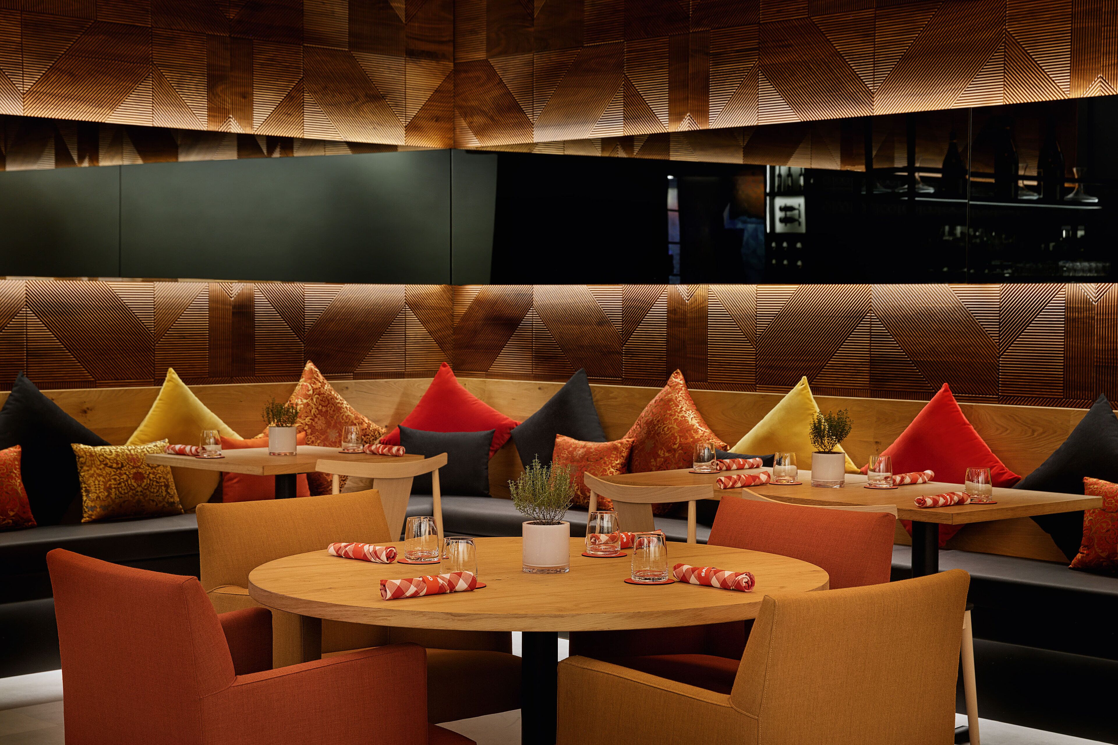 A modern restaurant with round tables, orange and red upholstered seating, and geometric patterned walls.
