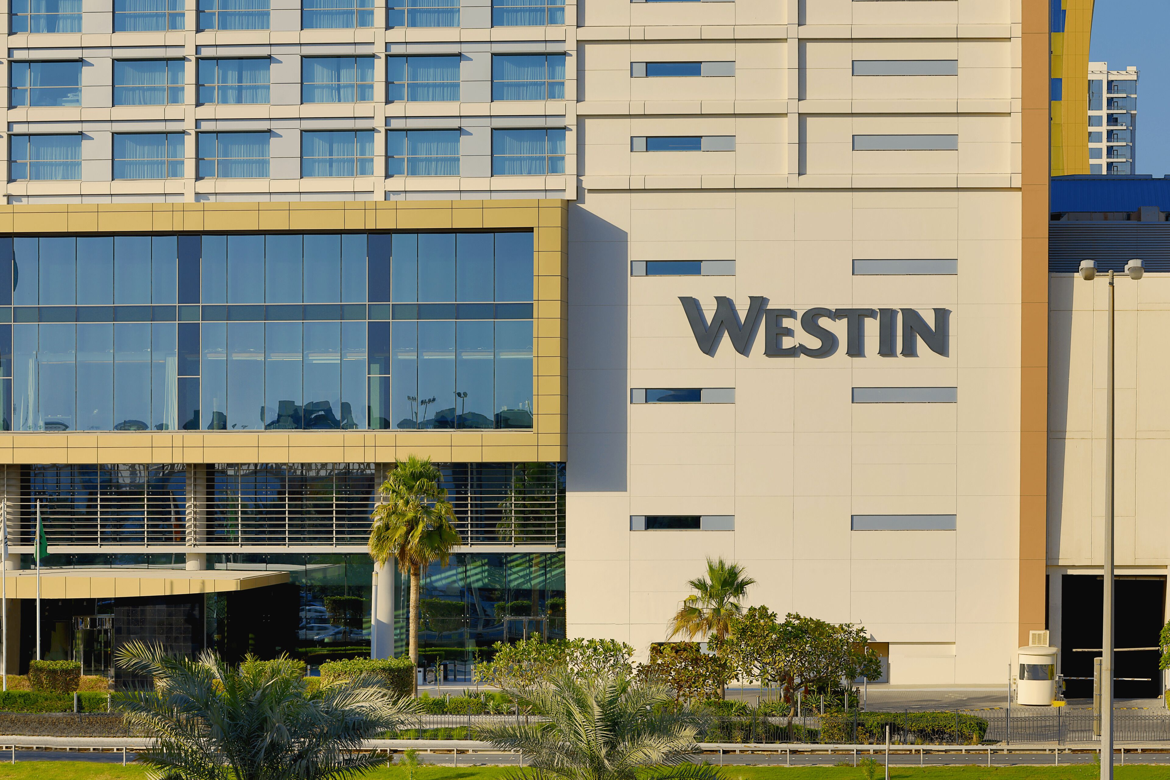 Un bâtiment moderne avec une grande enseigne « WESTIN » sur la façade, doté d’une entrée en verre et d’un espace paysager agrémenté de palmiers.
