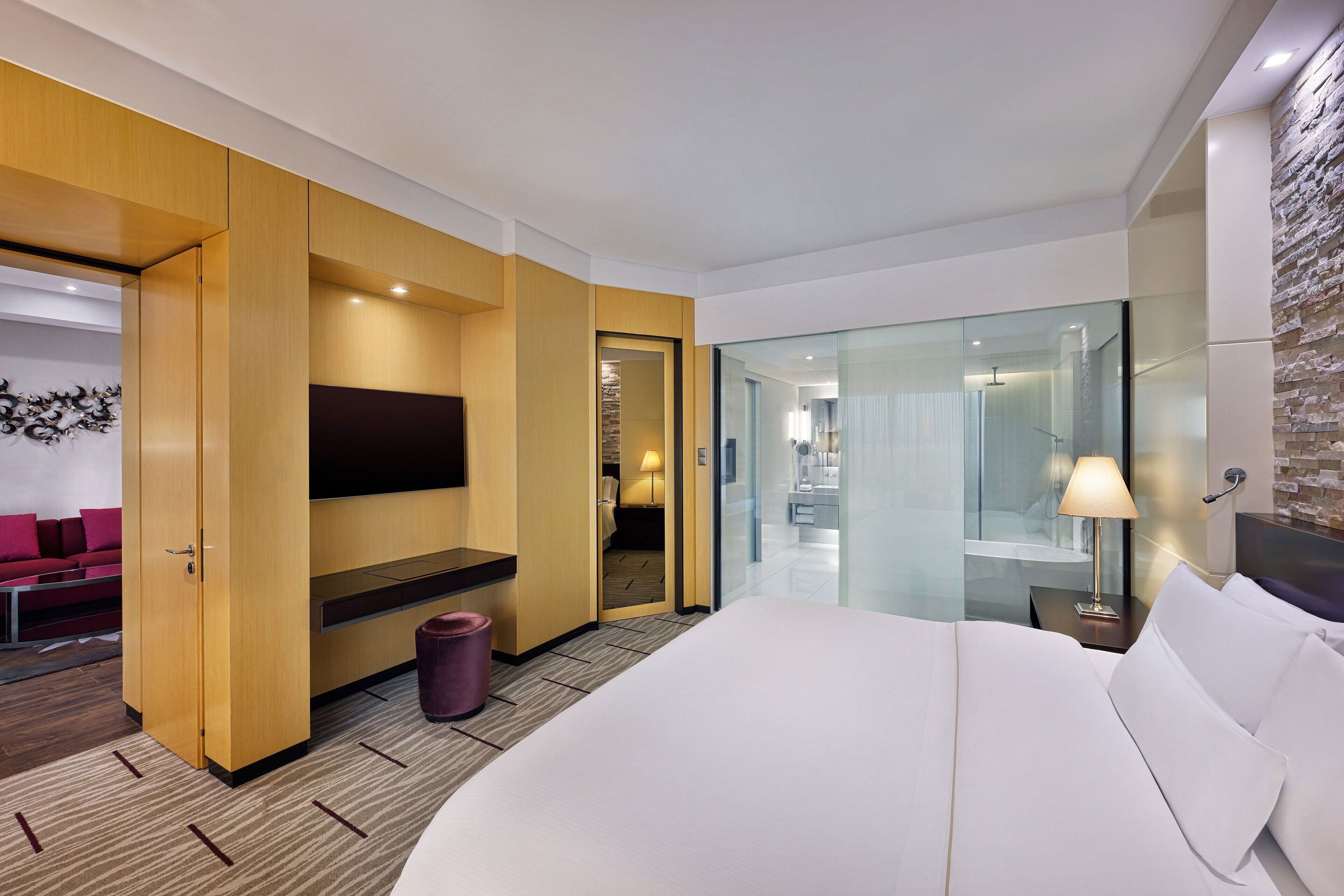 A modern hotel room with a large bed, a flat-screen TV mounted on the wall, a glass shower enclosure, and a wooden wardrobe.
