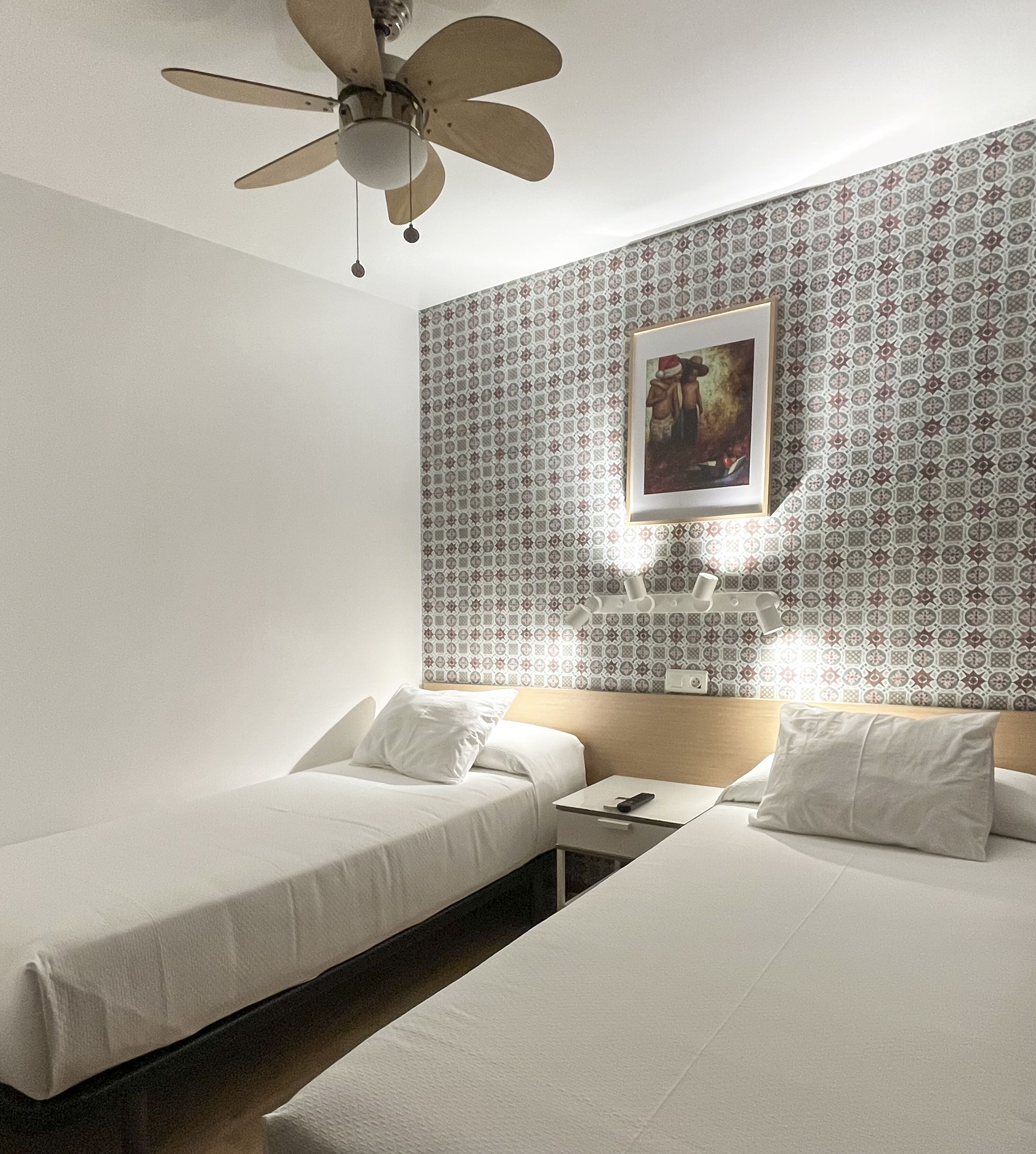 A hotel room with two single beds, a ceiling fan, patterned wallpaper, a framed picture on the wall, and bedside lamps.
