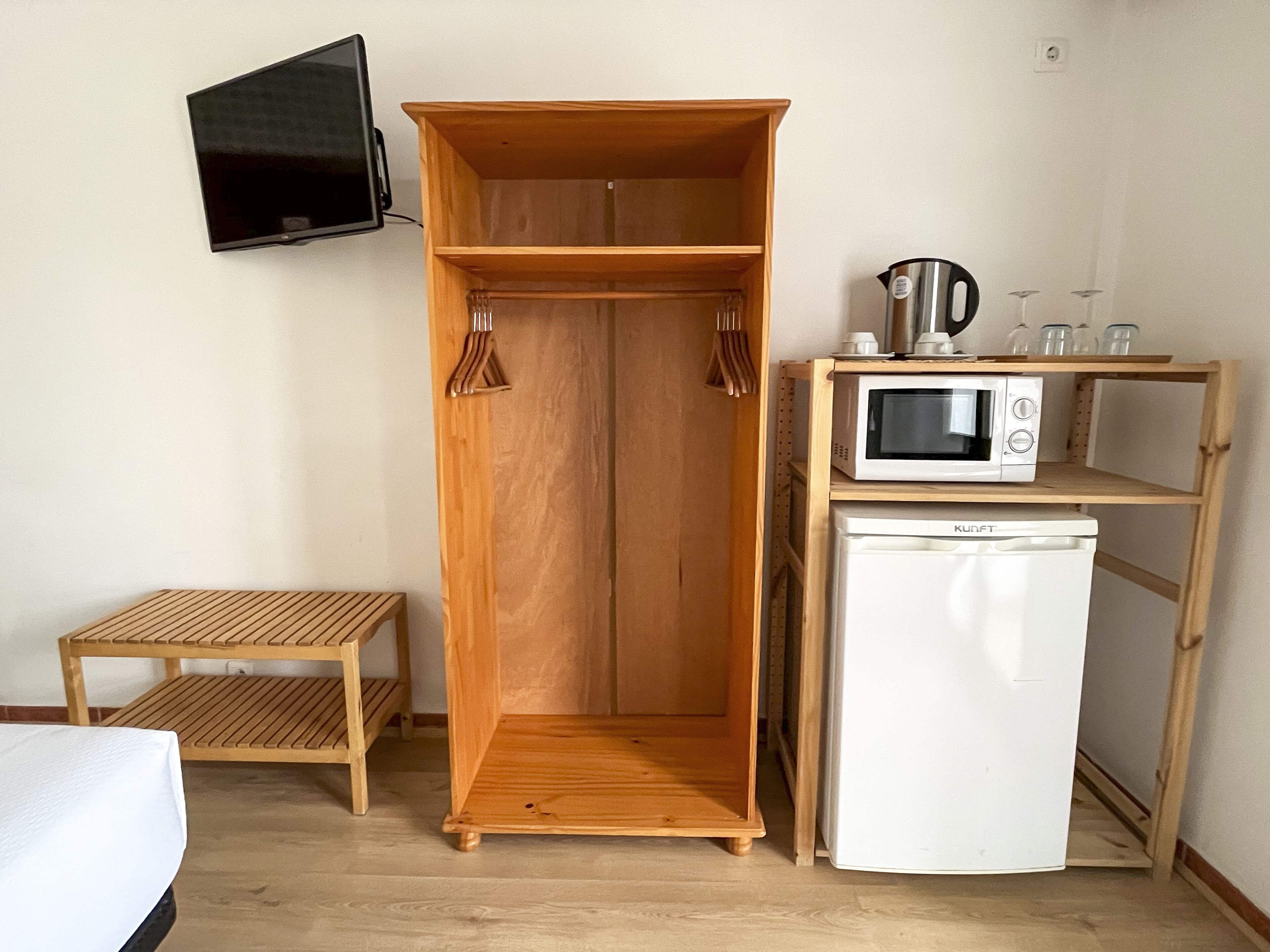 A compact room with a wooden wardrobe, a small wooden table, a white refrigerator, a microwave, and a television mounted on the wall.