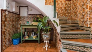 Una escalera con baldosas estampadas, un estante verde con libros y una planta en maceta, y una pared blanca con un dispositivo montado.
