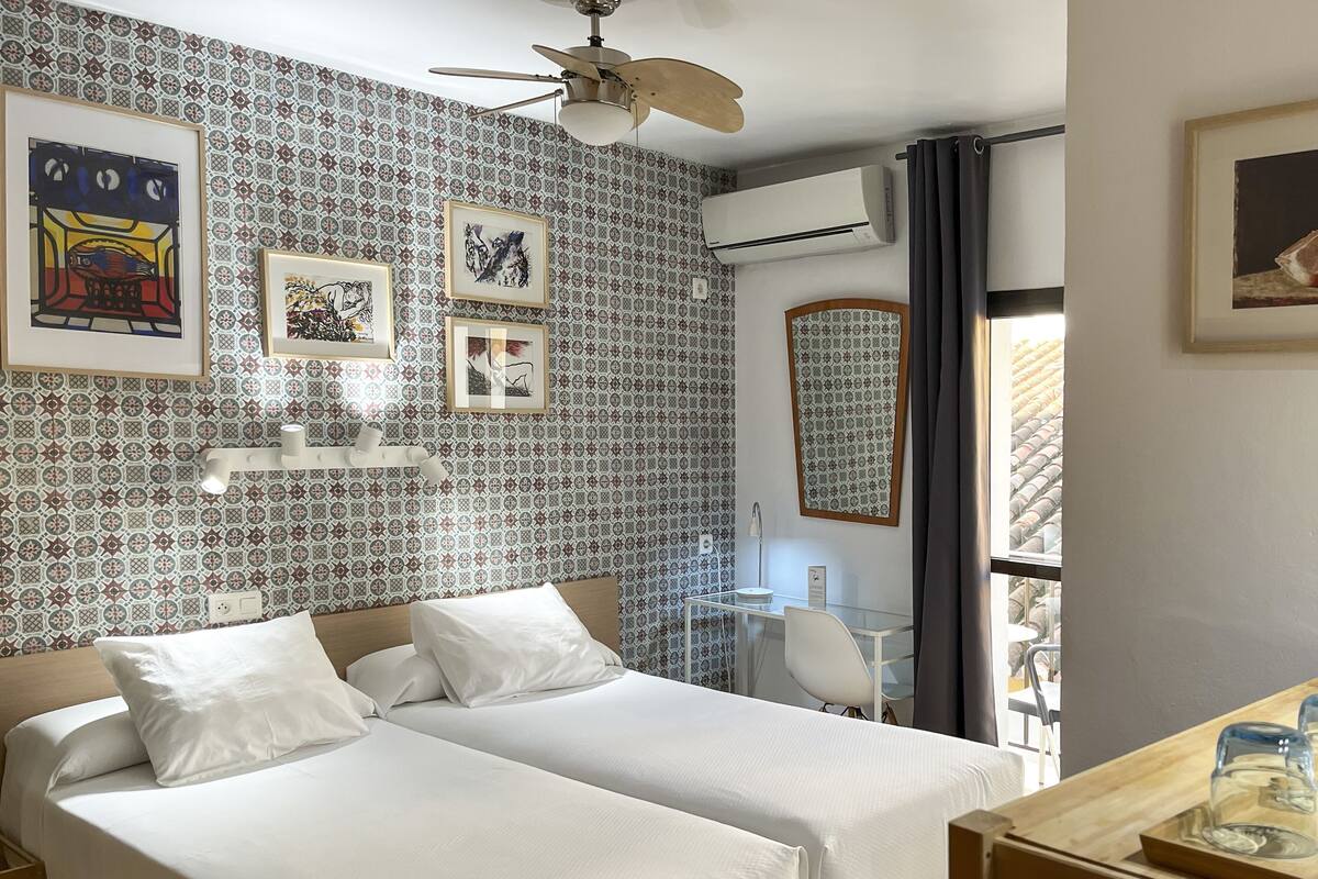Ein Hotelzimmer mit zwei Betten, einem Deckenventilator, strukturiertem Wandpapier, gerahmten Bildern, einem Fenster mit Vorhängen sowie einem Tisch mit Gläsern und einer Kiste.