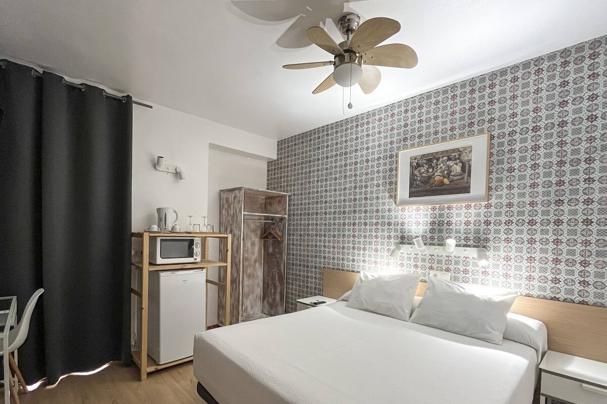 Ein Hotelzimmer mit Bett, Schreibtisch mit Stuhl, Kleiderschrank, Mikrowelle, Deckenventilator und einer Wand mit Muster.