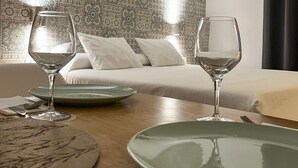 Un comedor ordenado con dos copas de vino, platos y cubiertos sobre una mesa de madera. La habitación presenta papel tapiz estampado, una cama con ropa de cama blanca y cuadros en la pared.