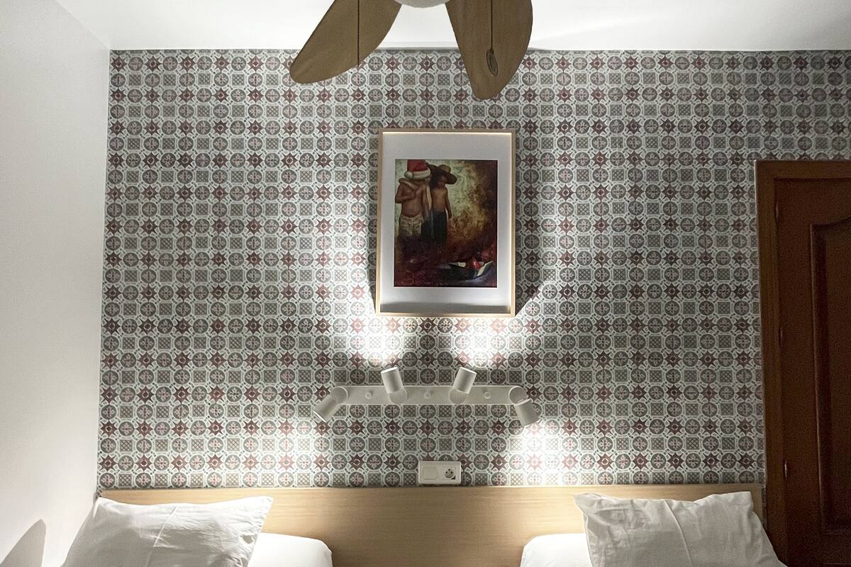 Ein Hotelzimmer mit zwei Betten, einem Deckenventilator, einem kleinen Tisch mit Fernbedienung und einer wandmontierten Lampe.