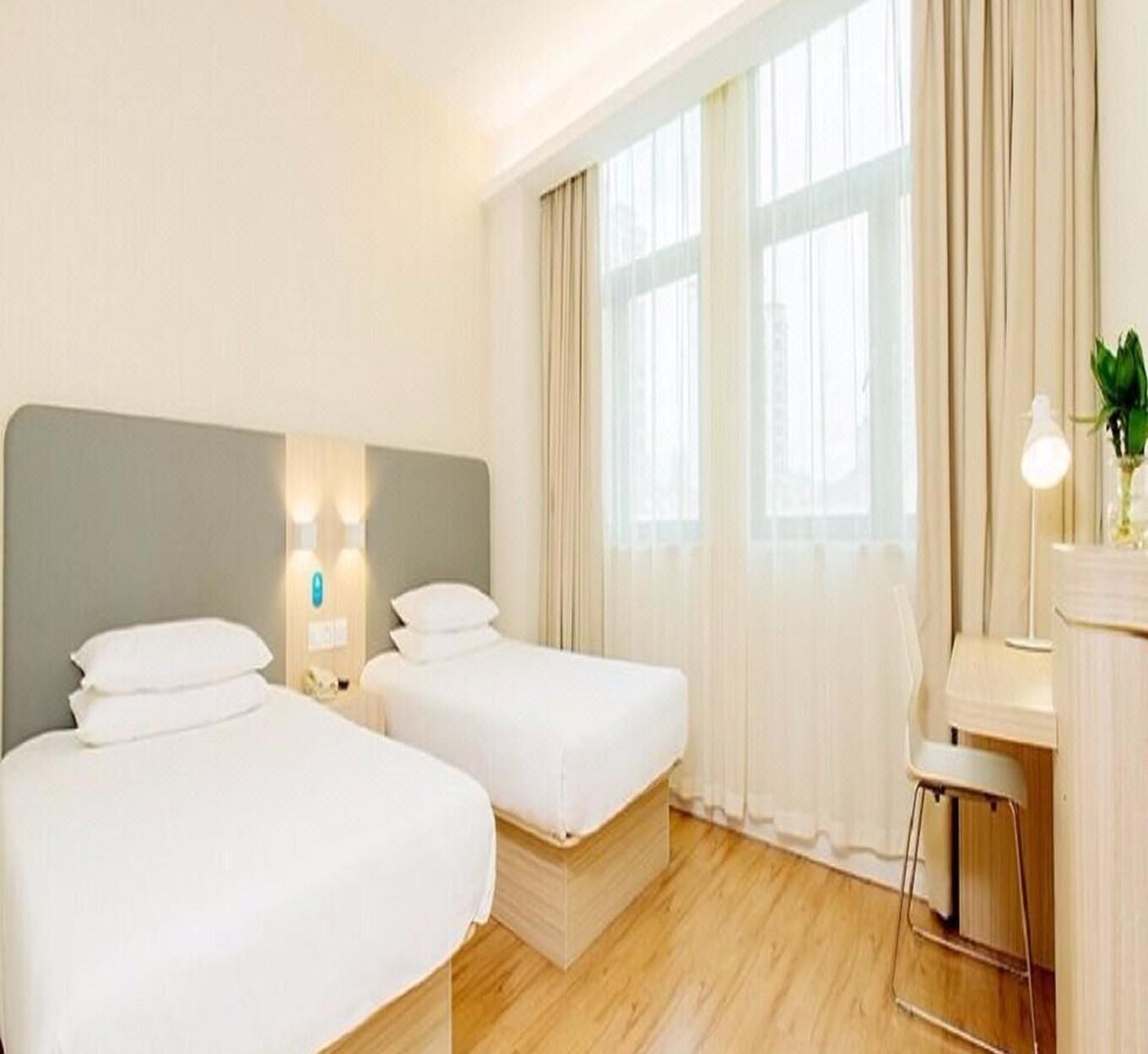 Une chambre d’hôtel avec deux lits simples, un bureau avec une chaise, une lampe et une grande fenêtre avec des rideaux.