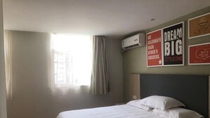 Habitación de hotel con cama, ventana con cortinas, equipo de aire acondicionado y carteles motivacionales en la pared.