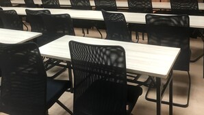 Un salón de conferencias con una larga mesa y múltiples sillas negras. Varias ventanas con persianas, una pizarra y una puerta al fondo.