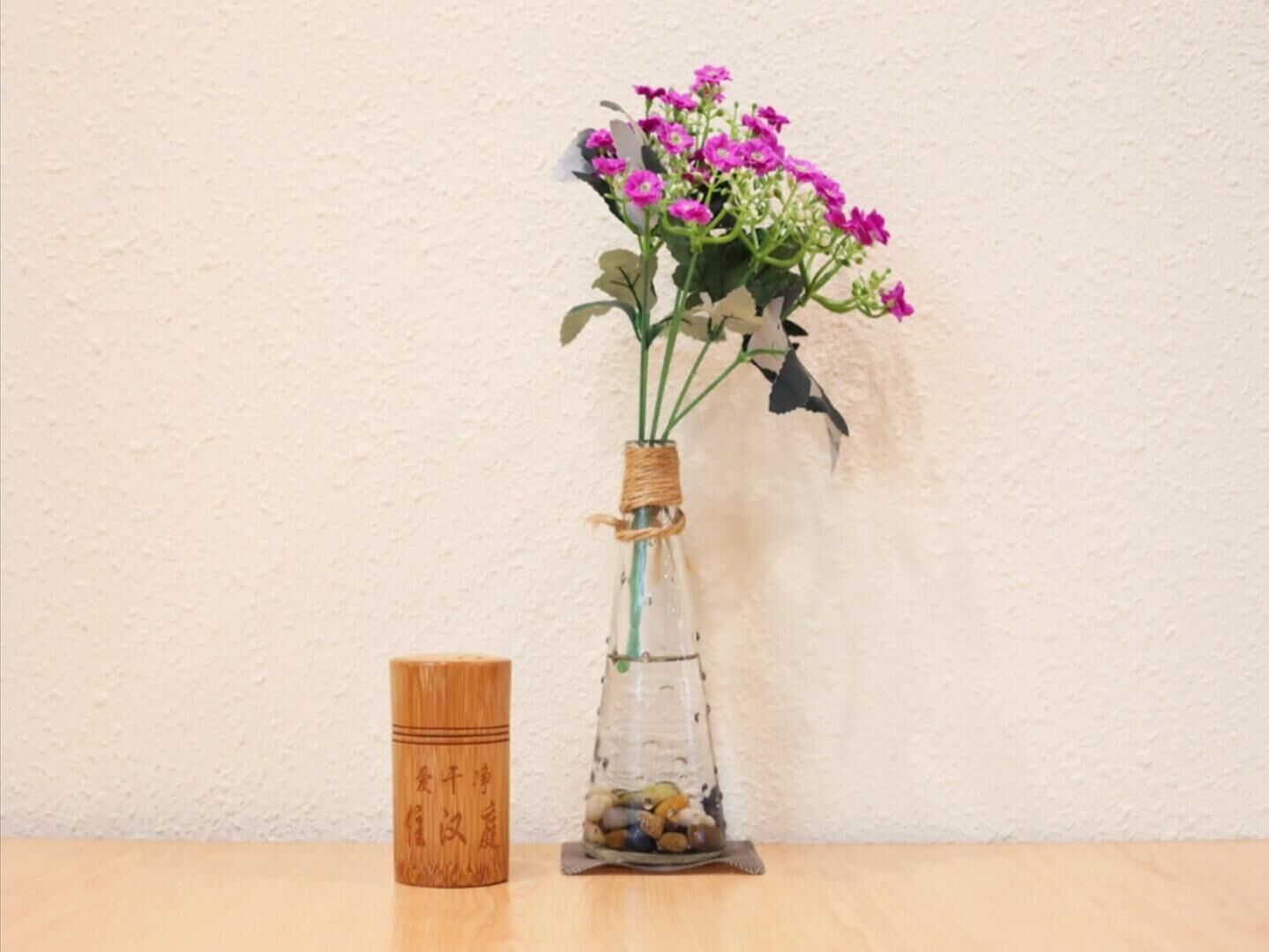 A clear glass vase with a bamboo base and a woven band, containing pink flowers, placed on a wooden surface against a plain wall.