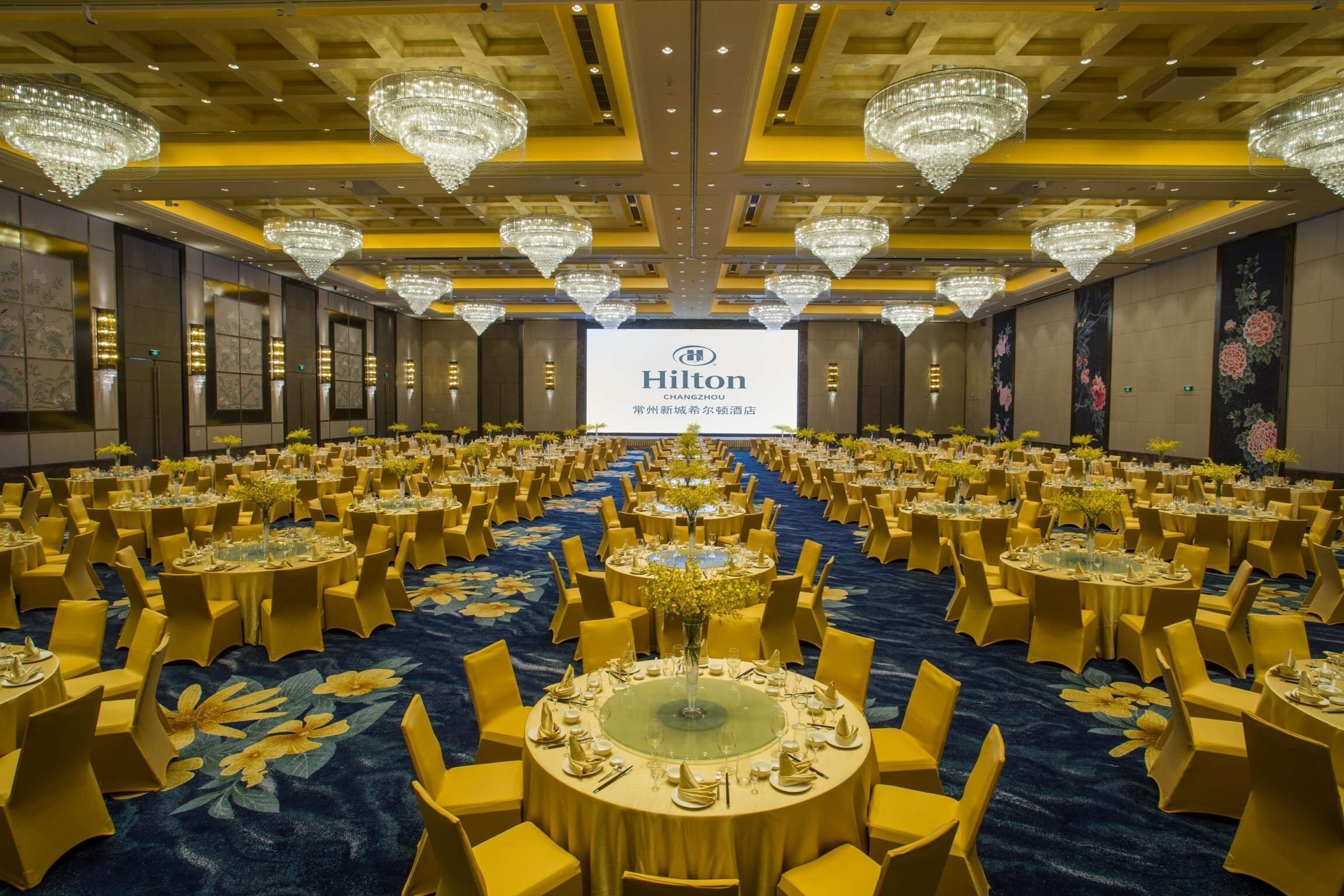 A spacious banquet hall with round tables, yellow chairs, and chandeliers.