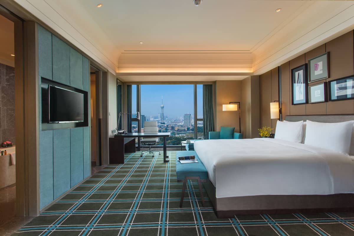 Une chambre d’hôtel avec un grand lit, un bureau, une chaise et une télévision. De grandes fenêtres offrent une vue sur la ville.